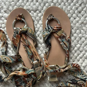Bohemiska sandaler med mönstrade band - Säljer ett par sandaler med platt sula och långa, bohemiskt mönstrade tygband i beige, brunt, blått och svart. Banden kan knytas runt vristen för en personlig look. Perfekta för varma dagar och festivaler. Storleken på sol är 36 men jag har 38 och passar mig. 