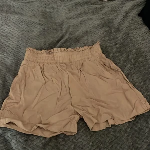 Beiga flowy shorts med resårmidja - Snygga beiga shorts med bred resår i midjan och volangkant upptill. De är luftiga och har en flowy passform, perfekta för varma dagar. Materialet känns mjukt och lätt, vilket gör dem riktigt sköna att bära. Är xs men mke stretch så passar S men tvek på M