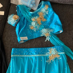 Dans dräckt - Superfin turkos tvådelad klänning från Patricia med glittriga pärlor och blommiga applikationer i guld och ljusblått. Toppen är ärmlös och kjolen är lång med transparent tyg och dekorativa detaljer. Perfekt för bal eller fest!