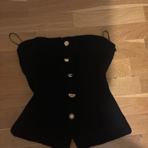 Svart korsettopp från Zara med knappar - Svart korsettopp från Zara med smala axelband och unika dekorativa knappar i olika former framtill. Toppen har en figurnära passform och är tillverkad i ett strukturerat tyg som ger en cool look. Perfekt för dig som vill sticka ut med detaljer.