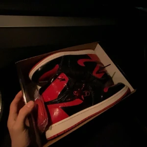 Jordan 1 Retro High OG Patent Bred - Säljers ett par Nike Air Jordan 1 Retro High OG Patent Bred med glansig finish i svart och rött. Klassisk high-top siluett med röd Swoosh, svarta snören och vit mellansula. Perfekta för dig som vill sticka ut med ikoniska sneakers. Authentic tag✅