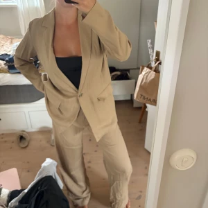 Beige kostym med kavaj och byxa - Säljer en snygg beige kostym med avslappnad passform. Kavajen har klassisk krage, knäppning framtill och fickor med lock. Byxorna är raka och vida i modellen. Perfekt för dig som gillar en stilren och trendig look. Storlek 40 i byxa och medium i kavaj 