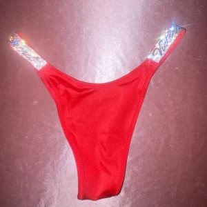Röda bikiniunderdelar Victoria's Secret - Röda bikiniunderdelar från Victoria's Secret med högt skuren modell och glittriga axelband med logga. Materialet är mjukt och stretchigt, huvudsakligen polyamid och elastan. Perfekt för dig som vill sticka ut på stranden med extra bling.