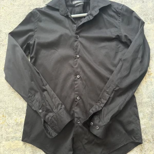 Svart slim fit skjorta Dressmann S - Säljer en svart skjorta från Dressmann i storlek S, tillverkad i stretchig bomull för en bekväm passform. Jag fick den i present av mina föräldrar men den passade tyvärr inte, och eftersom jag tagit bort etiketterna kan jag inte returnera den. Kvitto finns vid behov. Priset kan diskuteras 
