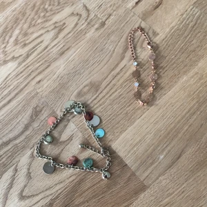 Två färgglada fotlänk med berlocker - Säljer två fotlänk: ett i guldfärgade metall med runda berlocker i turkos, röd, rosa och silver, samt ett i roséguld med små utskurna blommor och pärlliknande detaljer. Perfekta accessoarer för att liva upp din stil. Säljer båda för 10 kr