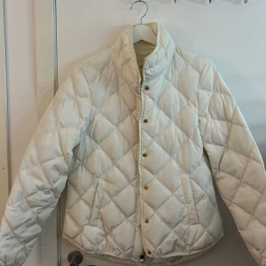 Vit quiltad jacka från Lindex - Säljer en stilren vit quiltad jacka från Lindex. Jackan har en hög krage och knäppning med tryckknappar. Perfekt för kyligare dagar med sin mysiga och varma design.