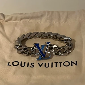 Louis Vuitton silverfärgat armband - Snyggt chunky armband från Louis Vuitton i blankt silverfärgat metall med kraftiga länkar och ikoniska LV-loggan i blått framtill. Perfekt statement piece för dig som gillar exklusiva accessoarer och vill sticka ut med en lyxig detalj.