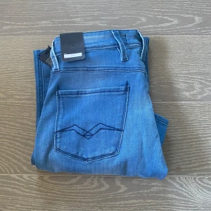 Blå Replay Anbass jeans slim fit - Snygga blå Replay Anbass jeans med klassisk femficksdesign och diskreta slitningar. Jeansen har slim fit-passform, normal midja och är tillverkade i stretchig bomullsdenim för extra komfort. Perfekta för dig som gillar en modern och stilren look.
