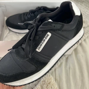 Svarta sneakers från Calvin Klein Jeans - Stilrena svarta sneakers från Calvin Klein Jeans med vita detaljer på hälen och logga på sidan. Skorna har snörning, rund tå och platt sula. Ovandelen är i textil och mocka för en snygg kontrast. Perfekta för en clean och modern look.