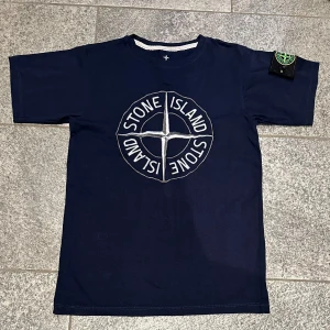  Stone Island t-shirt  - Snygg mörkblå t-shirt från Stone Island med stort broderat logomärke på bröstet och klassisk patch på ärmen. T-shirten är i bomull och har en normal passform med korta ärmar. Perfekt för dig som gillar streetwear och vill sticka ut med en ikonisk look.