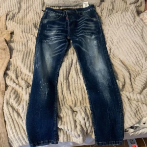 Dsquared2 blå jeans med slitningar - Snygga blå jeans från Dsquared2 med coola slitningar och färgstänk för en trendig look. Jeansen har knäppning med knappar, klassisk femficksmodell och smal passform. Perfekta för dig som gillar streetstyle och vill sticka ut lite extra. Aldrig använd (nya). Skinny jeans.