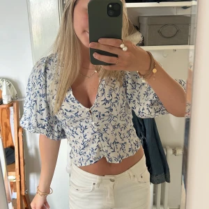 Blommig blus med puffärm och knappar - Superfin blus från H&M i vitt med blått blommönster. Modellen är croppad med v-ringning, puffiga korta ärmar och knappar framtill. 