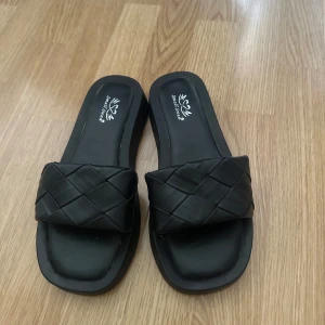 Svarta flätade sandaler - Svarta sandaler med bred, flätad rem i syntetmaterial och fyrkantig tå. Snygg minimalistisk design som passar perfekt till varma dagar. Platt sula för skön komfort och enkel att glida i. Passar Stl 39 
