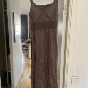 Lång brun klänning från CCDK - Snygg långklänning i brun nyans från CCDK med breda axelband och dragkedja bak. Klänningen har ett diskret blommigt mönster och markerad midja. Slutar nedanför knäna. 