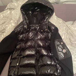 Moncler Cardigan - Snygg svart pufferjacka från Moncler med glansigt material och stickade ärmar. Jackan har en praktisk huva och dragkedja framtill. Har skada se bild 3