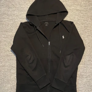 Svart hoodie från Polo Ralph Lauren - Säljer en stilren svart hoodie från Polo Ralph Lauren med dragkedja och det klassiska logotypbroderiet på bröstet. Perfekt för en avslappnad look. Den har långa ärmar och en praktisk huva med dragsko.