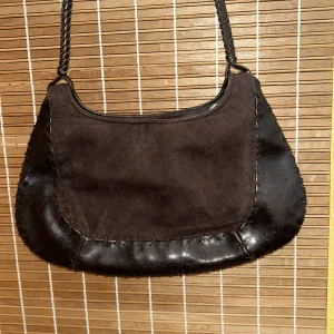 Brun axelväska i mocka och skinn - Snygg brun axelväska i en kombination av mocka och skinn. Väskan har en flätad axelrem och en praktisk dragkedja på baksidan för extra förvaring. 