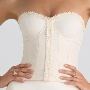 Krämfärgad korsett med spetsdetaljer - Elegant krämfärgad korsett med spetsdetaljer längs kanten och snörning i ryggen. Den har en axelbandslös design och ger en smickrande passform. Perfekt för att skapa en stilren och sofistikerad look.
