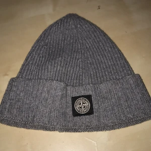 Grå ribbad mössa från Stone Island - Snygg grå ribbad mössa från Stone Island med deras ikoniska logga framtill. Perfekt för att hålla värmen under kyliga dagar. Stilren design som passar till det mesta.