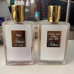 Parfymflaskor från By Kilian - Två eleganta parfymflaskor från By Kilian med guldfärgade lock och etiketter. Den ena flaskan är märkt 'Love, don't be shy' och den andra 'Voulez-vous coucher avec Moi'. Flaskorna har en stilren och lyxig design med genomskinligt glas som visar vätskan inuti. 
