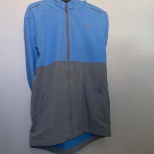 Blå och grå jacka från Under Armour - Snygg blå och grå jacka från Under Armour med dragkedja och huva. Perfekt för träning eller en avslappnad dag. Jackan har praktiska fickor med dragkedja framtill. Xs men passar S för den är lite oversized 