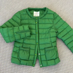 Grön dunjacka från Holly & Whyte - Säljer en snygg grön dunjacka från Holly & Whyte by Lindex. Jackan har långa ärmar och stängs med knappar framtill. Den har praktiska fickor och en quiltad design som ger en stilren look. Perfekt för kyligare dagar!