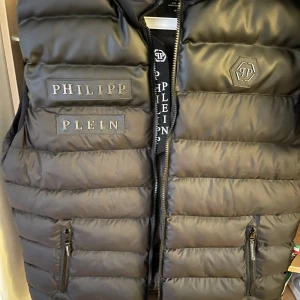 Svart dunväst från Philipp Plein - Säljer en svart dunväst från Philipp Plein med dragkedja och logotypdetaljer på bröstet. 