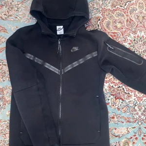 Svart Nike tech  - Säljer en svart hoodie från Nike med dragkedja och huva. Jackan har en diskret logga på bröstet och en praktisk ficka med dragkedja på ärmen. Perfekt för en sportig look eller avslappnade dagar.
