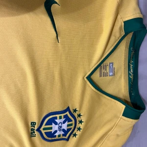 Gul Brasilien fotbollströja från Nike - Säljer en gul Brasilien fotbollströja från Nike med gröna detaljer och korta ärmar. Tröjan har Brasiliens emblem på bröstet och en Nike-logga. Perfekt för fotbollsfans som vill visa sitt stöd för laget!