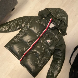 Grön pufferjacka från Moncler - Säljer en snygg grön pufferjacka från Moncler med huva och dragkedja. Jackan har en ikonisk röd, vit och blå rand längs dragkedjan och en broderad logotyp på bröstet. Perfekt för kyliga dagar!