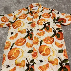 Färgglad kortärmad skjorta med apelsinmönster från Vailent - Säljer en färgglad kortärmad skjorta från Vailent med ett livligt apelsinmönster. Skjortan har knappar framtill och är perfekt för en somrig look. Materialet är lätt och bekvämt.