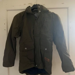 Grön jacka från KappAhl - Säljer en grön jacka från KappAhl med huva och knappar. Jackan har en klassisk design med praktiska fickor och är perfekt för kyligare dagar.