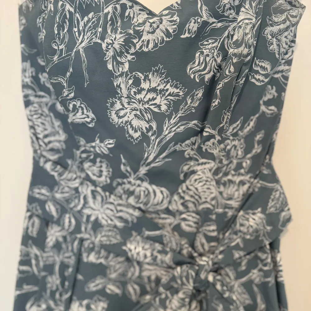 Elegant klänning i en dämpad blå nyans med vackert blommönster i vitt. Klänningen har en v-ringad hals och ett knytband i midjan som ger en smickrande passform. Perfekt för sommarens festligheter.. Mekot.