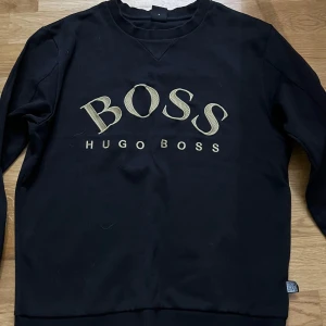Hugo Boss tröja - Snygg svart tröja från Hugo Boss med guldtryck på bröstet. Tröjan har långa ärmar och en rund halsringning. Perfekt för en stilren look.