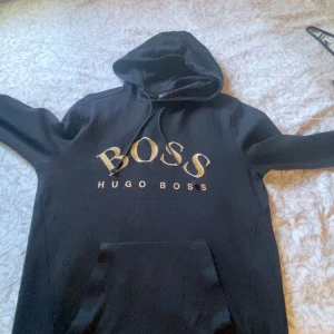 Svart hoodie från Hugo Boss - Säljer en stilren svart hoodie från Hugo Boss med guldtryck på bröstet. Tröjan har en praktisk dragkedja vid ärmslutet och en stor ficka framtill. Perfekt för en avslappnad och trendig look. Säljer till bra pris.  den är slutsåld överallt. Dunder kvalité! Det är storlek s men sitter som m 