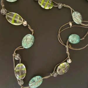 Halsband med gröna och turkosa pärlor - Halsband med stora, ovala pärlor i grönt och turkos. Pärlorna är genomskinliga med inslag av gröna ränder och har dekorativa metallrosor mellan dem. Perfekt för att ge en färgklick till din outfit!
