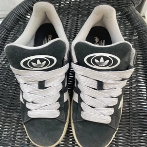 Svarta Adidas Campus sneakers - Snygga svarta Adidas Campus sneakers med vita detaljer och klassiska tre ränder på sidan. Skorna har en bekväm gummisula i ljusbrun färg och är tillverkade i mocka. Perfekta för en stilren och avslappnad look.