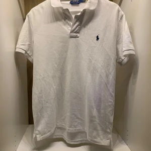 Vit pikétröja från Ralph Lauren - Säljer en klassisk vit pikétröja från Ralph Lauren i custom slim fit. Tröjan har en broderad logga på bröstet och korta ärmar. Perfekt för en stilren look! En knapp har lossnat men enkelt o fixa!
