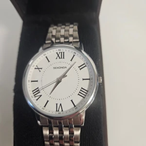 Klassisk klocka från Sekonda - Elegant klocka från Sekonda med en stilren vit urtavla och romerska siffror. Armbandet är i silverfärgad metall och har en klassisk länkdesign. Perfekt för en tidlös look.