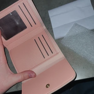 Liten plånbok - Säljer en stilren rosa liten plånbok. Plånboken har flera kortfack och en knappstängning. Perfekt för att hålla ordning på dina kort och kontanter.