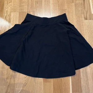 Svart kortkjol - Snygg svart kortkjol med en enkel och stilren design. Perfekt för att matcha med olika toppar och accessoarer. Kjolen har en lätt utsvängd form som ger en fin siluett.