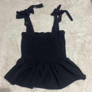 Svart topp med knytband 🖤 - Säljer en svart topp från Shein med smockad design och knytband på axlarna. Toppen har en volangdetalj vid nederkanten som ger en söt touch. Perfekt för sommarens varma dagar! 🖤