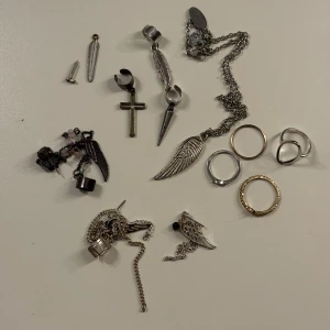 Smyckeset med örhängen, ringar och armband - Ett varierat smyckeset med flera örhängen och ringar. Örhängen inkluderar motiv som kors och vingar, samt detaljer med kedjor och svarta pärlor. Ringarna är i både silver och guld, med olika former och mönster. Armbanden är i varierande turkosfärgade stenar. 