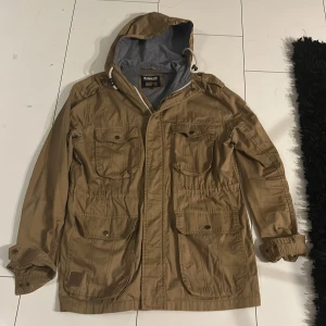 Field jacket från Bondelid - Säljer en beige field jacka från Bondelid med huva och flera praktiska fickor. Jackan har dragkedja och knappar framtill samt justerbara ärmslut. Perfekt för vår och höst!