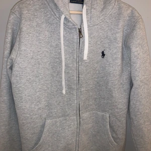 Grå Ralph Lauren zip up - Säljer en stilren grå zip up från Ralph Lauren med dragkedja och vit snörning i huvan. Den är nyskick och säljer pågrund av att den M blev för stort för mig. Ställ gärna frågor om pris eller annat.