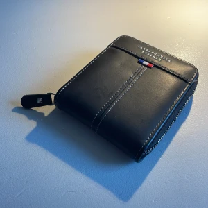 Läder plånbok från Frankrike  - Snygg svart plånbok i skinn från Carrken Style. Den har en praktisk dragkedja och flera fack för kort. En liten detalj i rött, vitt och blått ger en stilren touch. Perfekt för att hålla ordning på dina kort och kontanter. Plånbok från Frankrike 