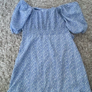 Blå blommig klänning - Kom privat för mer bilder, Charmig blå klänning med blommigt mönster och puffiga kortärmar. Perfekt för en somrig look med sin offshoulder-design.