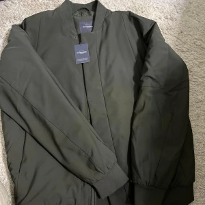 Grön bomberjacka från Dressmann - Snygg grön bomberjacka från Dressmann i storlek XL. Jackan har en dragkedja framtill och ribbade muddar vid ärmslut och nederkant. Perfekt för en stilren look. Nypris: 799kr. Helt ny, aldrig använd.