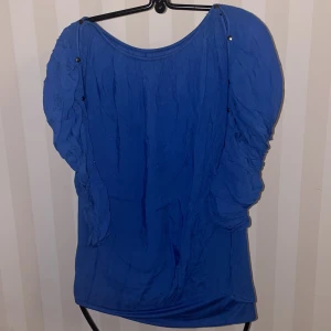 Blå topp med detaljerade ärmar st.S-M - Snygg blå topp med unika detaljer på ärmarna. Toppen har en rund halsringning och dekorativa knappar längs ärmarna som ger en extra touch. Perfekt för att piffa upp din garderob!