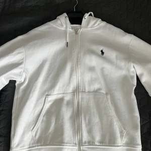Vit hoodie från Polo Ralph Lauren - Säljer en stilren vit hoodie från Polo Ralph Lauren med dragkedja och det klassiska logotypbroderiet på bröstet. Tröjan har en bekväm passform med långa ärmar och praktiska fickor framtill. Perfekt för en avslappnad look.
